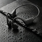 Black sport Audionex Bone Conduction Headphones with a continuous headband, shown with water droplets on a dark slate surface.