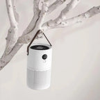 A white cylindrical portable SkyPhis Air Purifier with a dark brown hanging strap, suspended from a white tree branch against a light off-white background.