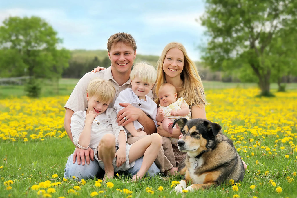 A family of five and a dog pose together in a grassy field filled with yellow flowers. The father and mother sit behind their two children, who are holding a baby, while a black, tan, and white dog lies in the foreground.