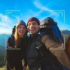 A smiling couple takes a selfie while hiking in a mountain landscape under a bright blue sky. Both are wearing large hiking backpacks and winter outerwear.