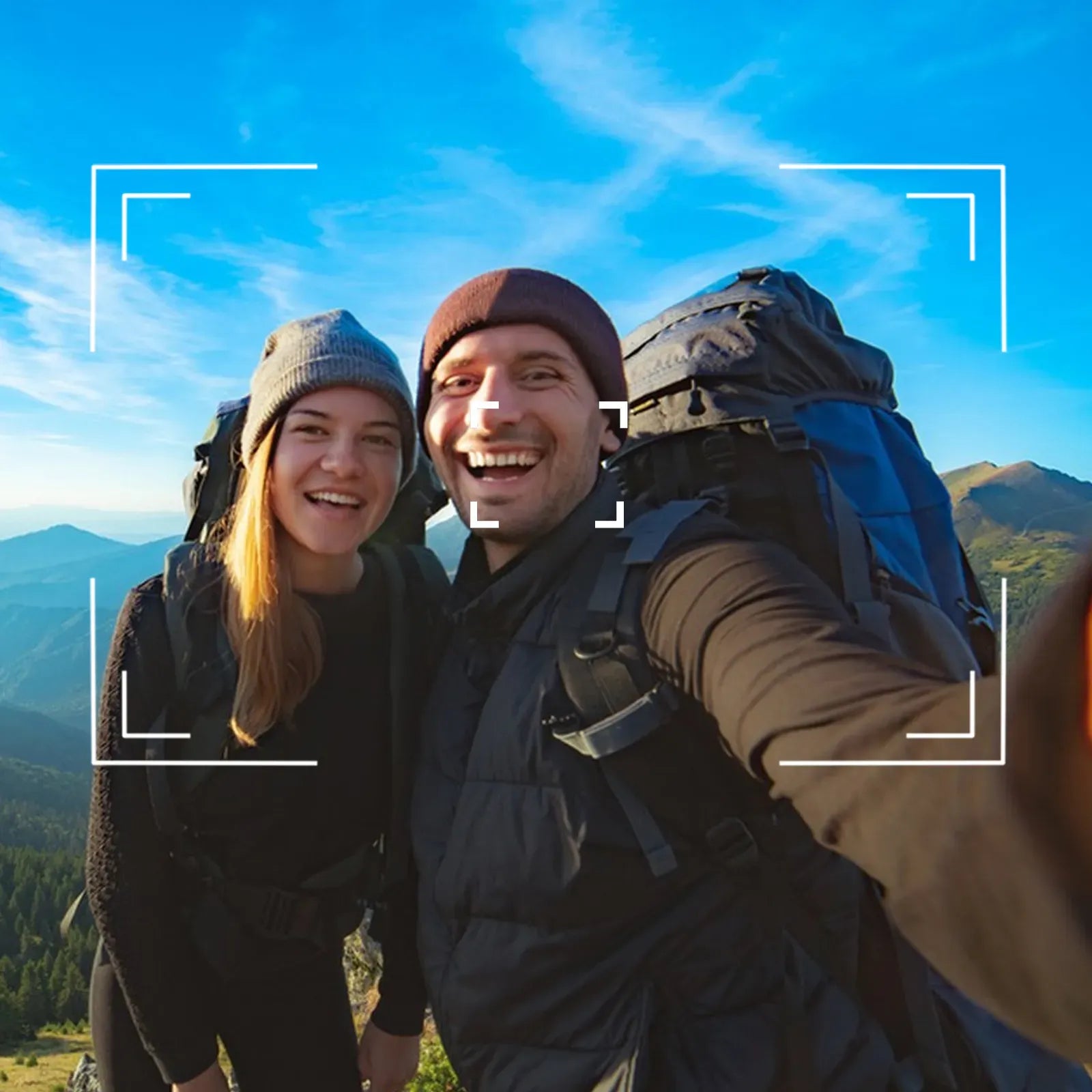 A smiling couple takes a selfie while hiking in a mountain landscape under a bright blue sky. Both are wearing large hiking backpacks and winter outerwear.